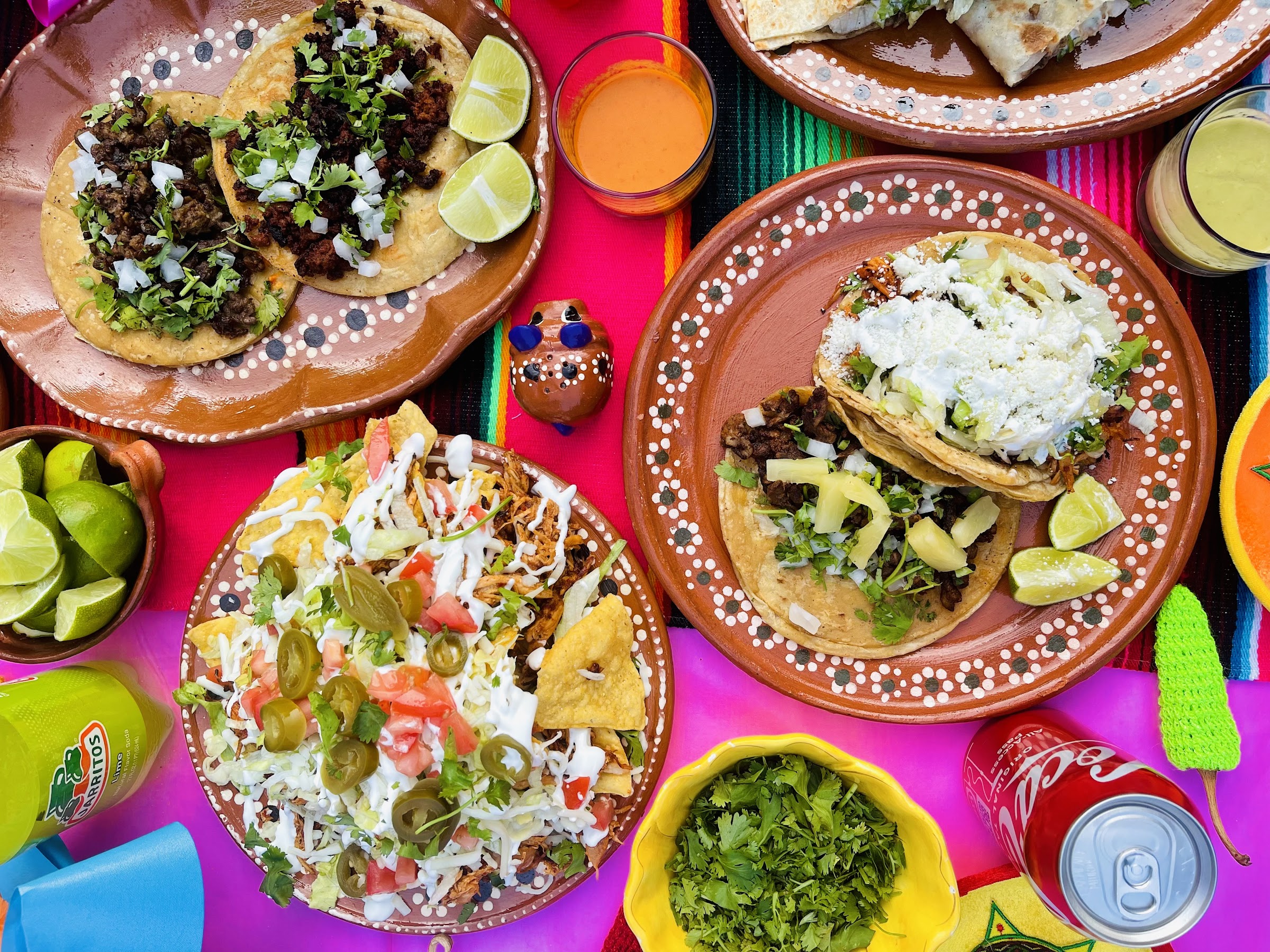 Overhead spread of tacos, loaded nachos and salsas on clay plates over a hot-pink serape-runner table at Taquero Mucho